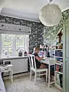 Ett rum med blommig tapet, stort fönster, vitt skrivbord och en person som sitter vid datorn.