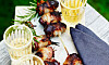 Grillspett med dadlar och bacon på en bricka bredvid glas och en mörk servett.