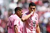 Fotbollsspelare i rosa tröja håller fingret mot munnen med en lagkamrat i bakgrunden.