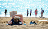 En person ligger på sanden och läser en bok på en strand med flera badgäster i bakgrunden.