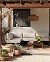 En uteplats med beige soffa, trätbord, terrakottakrukor och växter framför en putsad vägg.