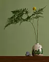En keramisk vas med grön Mumin-dekor står på ett träbord bredvid kvistar och en liten stenfigur mot grön bakgrund.