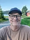 En man med keps och glasögon tar en selfie utomhus på en grusväg vid sjö och skog.