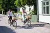 Två personer cyklar på en gata med vita hus, gröna staket och träd i solsken.