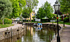 En smal kanal med små båtar, trädkantade gångvägar och en gatlykta på en grönskande sommardag.