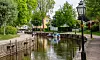 En smal kanal med små båtar, trädkantade gångvägar och en gatlykta på en grönskande sommardag.