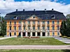 Ett stort gulbeige slott med mörkt tak och gräsmatta framför under en blå himmel.