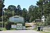 En vit byggnad med gröna knutar vid en grind, flaggstång, ett till vitt hus och skog i bakgrunden.