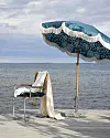 Blårandigt parasoll med fransar bredvid en stol med ljus filt på en brygga vid havet.