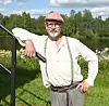 En man med keps och glasögon står vid ett räcke nära en älv omgiven av grönska.