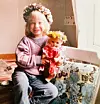 Ett barn sitter vid ett fönster och håller en docka med blomsterkrans på en blommig soffa.