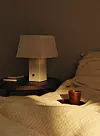 En bordslampa med ljus skärm står på ett nattduksbord intill en säng med beige täcke och en kopp.