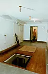 Ett sparsamt inrett rum med en öppen lucka i golvet, en matta och en person vid dörröppningen.