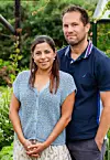 Två personer står framför en grön och blommande trädgård med buskar och växter i bakgrunden.