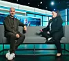 Två personer sitter i en soffa i en upplyst tv-studio med en stor skärm bakom sig.