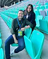 Två personer sitter på turkosa stolar i en tom fotbollsarena.