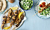 Grillspett, citronklyftor, sallad och tallrikar med mat på ett blått bord.