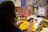 En person sitter vid ett bord med broderimaterial, garnnystan och små porslinsfigurer i en ateljé.