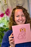 Kvinna ler och visar en rosa broderad tavla med en hund.