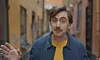 En man med mustasch står i en smal gränd och gestikulerar mot kameran.