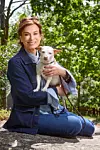 En kvinna sitter på en sten med en liten vit hund i knät framför träd och buskar.