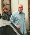 Två personer står vid en bil utanför en ljus byggnad.