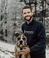 En leende man i svart tröja håller en brunvit hund i sele utomhus bland snötäckta träd.