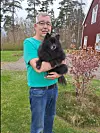 En man i turkos T-shirt håller en svart liten hund utomhus framför ett rött hus.