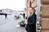 En kvinna står vid en byggnad på en kullerstensgata med cyklar och förbipasserande i bakgrunden.