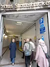 Flera personer går in i en garageinfart med blå parkeringstavla och väggmonterade skyltar.