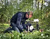 En person knäböjer i en skogsglänta och justerar kamerautrustning bland grönt gräs och vita blommor.