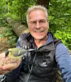 En man i väst sitter i skogen och matar en liten fågel ur handen.