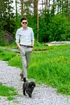 En man i ljus skjorta promenerar med en liten svart hund på en grusväg i en grön lantlig miljö.