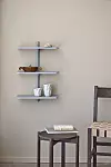 Väggmonterad hylla med små keramikföremål, ett sidobord och del av en stol mot beige vägg.