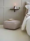 En beige puff står vid en väggmonterad hylla med ljus, vas och bok i ett sovrum.