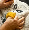 Händer broderar med gul och mörk ulltråd på ett ljust textilstycke.