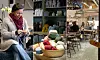 En kvinna stickar i en butik med garnhärvor, hyllor med keramik och ett cafélikt sittområde i bakgrunden.