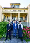 Gulmålat hotell med skylten Walaker Hotell och fem personer stående framför entrén bland gröna buskar och rosa blommor.