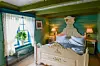Ett hotellrum med vit säng, gröna väggar, två sänglampor och ett fönster med vita spetsgardiner.