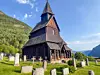 En mörk träkyrka omgiven av gravstenar på en grön sluttning med fjäll och fjord i bakgrunden.