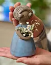 Händer håller en blå keramikfigur med ett brunt huvud och små vita blommor i handen.