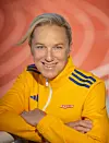 Porträtt av en person i gul träningsjacka med korsade armar mot orange bakgrund.