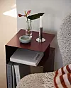 Liten mörk hylla med böcker, vas med blomma, skål och lampfot bredvid en fåtölj.