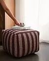 En hand placerar en kopp på en bok ovanpå en randig sittpuff vid ett fönster.