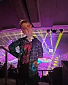 Person med glasögon står framför färgstarka laserljus i en fullsatt konsertarena.