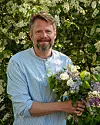En leende man i randig skjorta håller en bukett blommor framför blommande buskar.