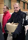 Två män utanför en byggnad i snöfall, den ena bär en kartong och står bredvid en mikrofon.