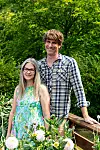 Två personer står på en träbro omgivna av grönska och blommor i en trädgård.
