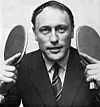 Svartvit bild av en man i kostym som håller två bordtennisracketar vid huvudet.