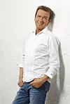 En man i vit skjorta och jeans står lutad mot en ljus vägg och tittar mot kameran.